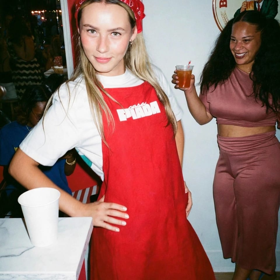 Two women pose inside a lively restaurant, one wearing a red pizza apron and the other holding a drink beneath a beer poster.