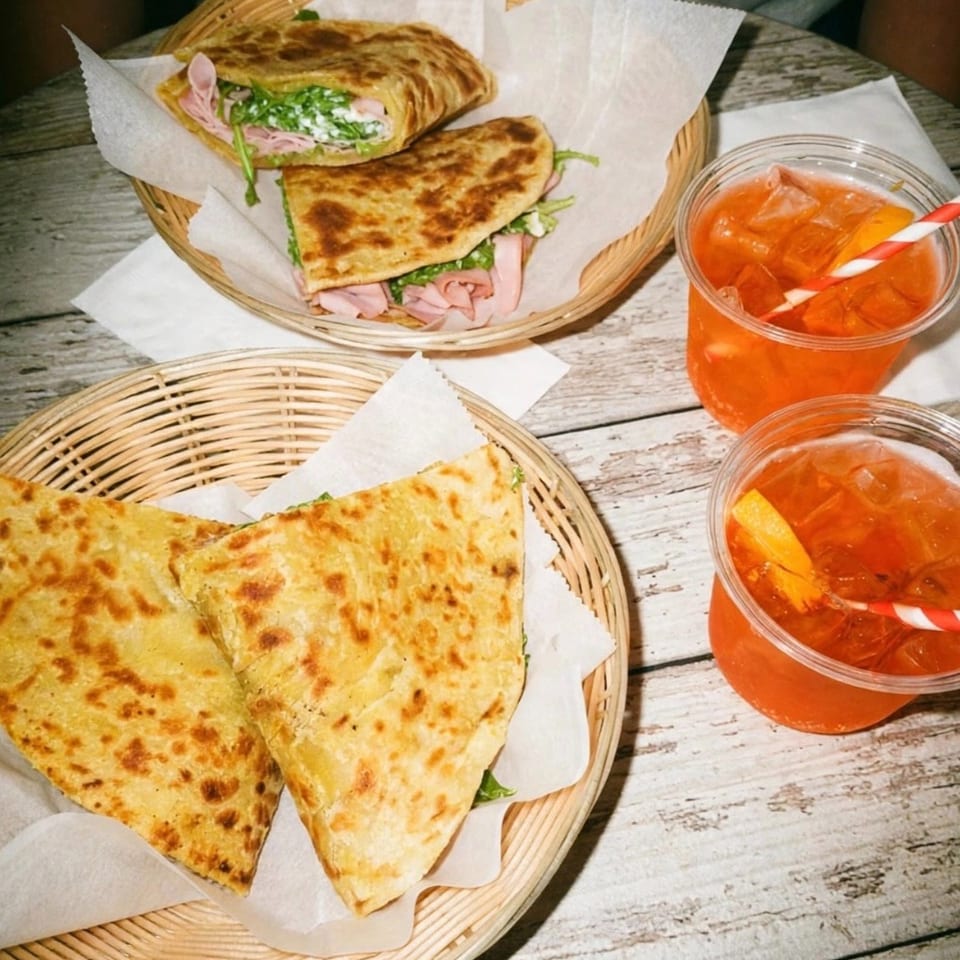 Wicker baskets hold toasted stuffed flatbreads with greens and sliced meat beside two orange iced drinks on a rustic table.