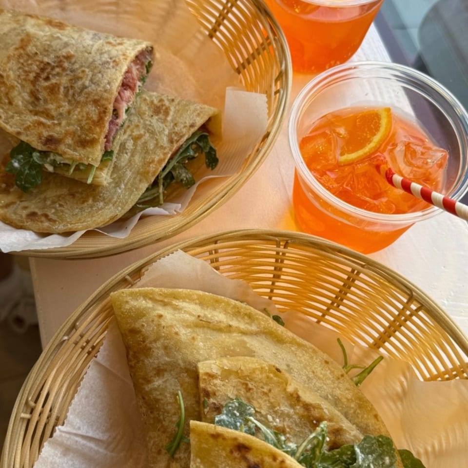 Baskets of toasted flatbread sandwiches with greens and meat beside two orange iced drinks garnished with lemon slices.