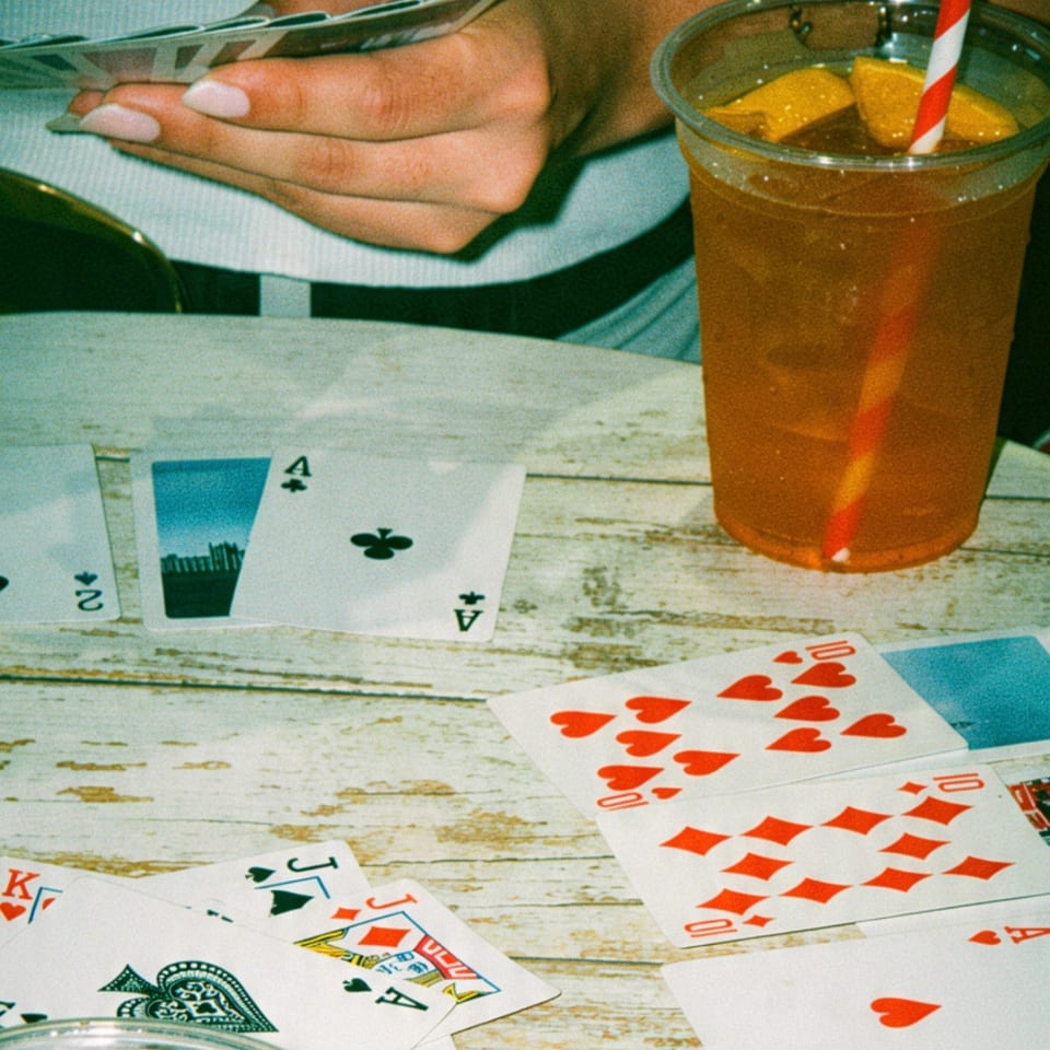 Playing cards spread across a wooden table beside iced drinks, a hand holding cards, and a red-and-white striped straw.