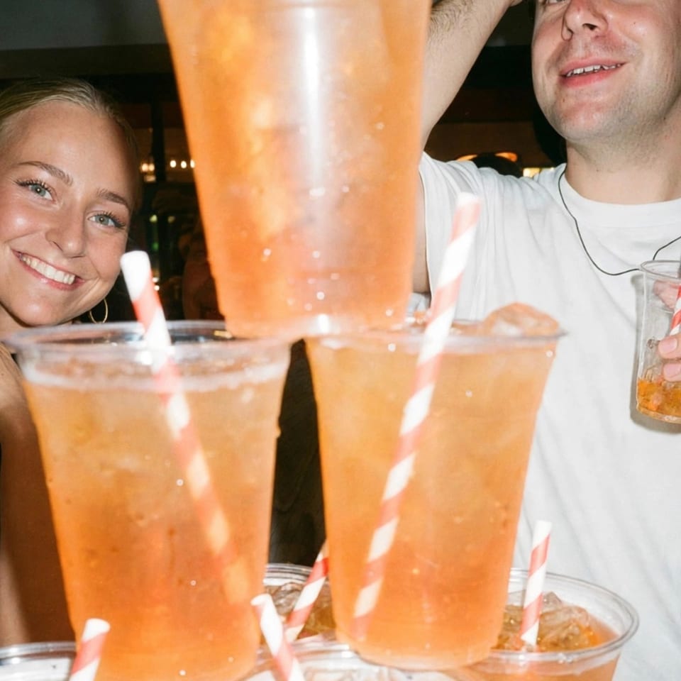 Two smiling people pose behind multiple orange drinks in clear cups with striped straws at a dimly lit restaurant.