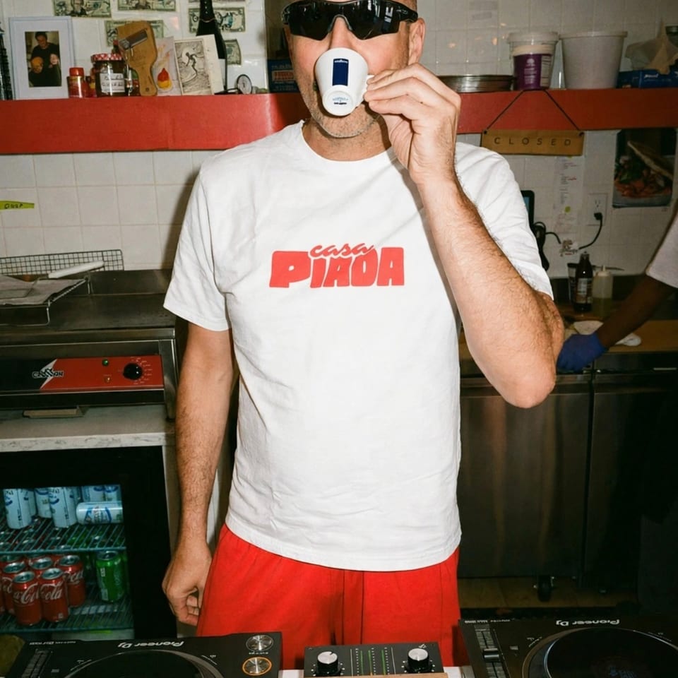 Bald man wearing sunglasses and a Casa Piasa shirt drinks from a cup behind a DJ setup in a restaurant kitchen.