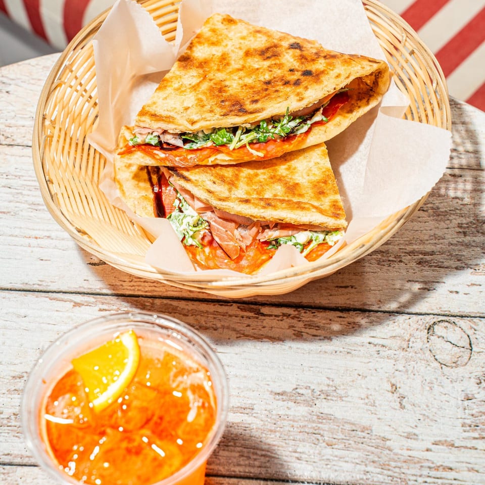 Grilled stuffed flatbread sandwiches with greens and sauce in a wicker basket, alongside an orange drink on a rustic table.