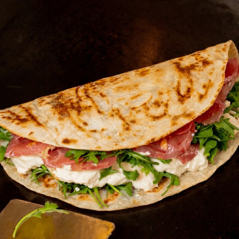 Grilled folded flatbread filled with creamy cheese, sliced cured meat, and fresh arugula on a dark cooking surface.
